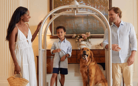 A smiling family of three and their golden retriever standing by a silver hotel luggage cart in a beautifully decorated lobby.