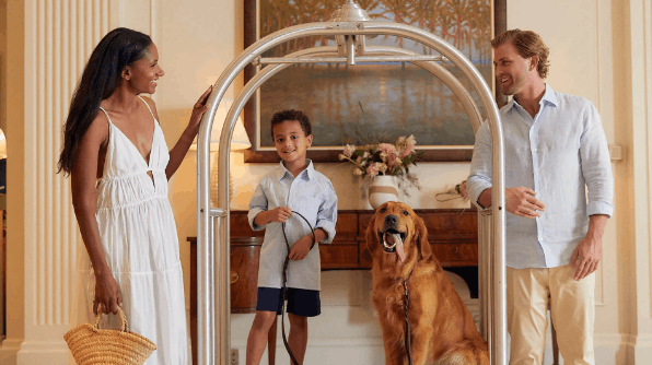 A smiling family of three and their golden retriever standing by a silver hotel luggage cart in a beautifully decorated lobby.