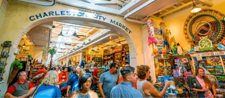Interior view of the bustling Charleston City Market, showing visitors browsing various vendor stalls under a high arched ceiling with hanging lights and fans.