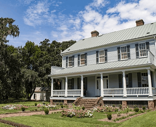 Historic McLeod Plantation house with a large front porch and a blooming garden under a blue sky.