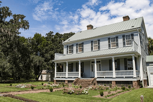 Historic McLeod Plantation house with a large front porch and a blooming garden under a blue sky.